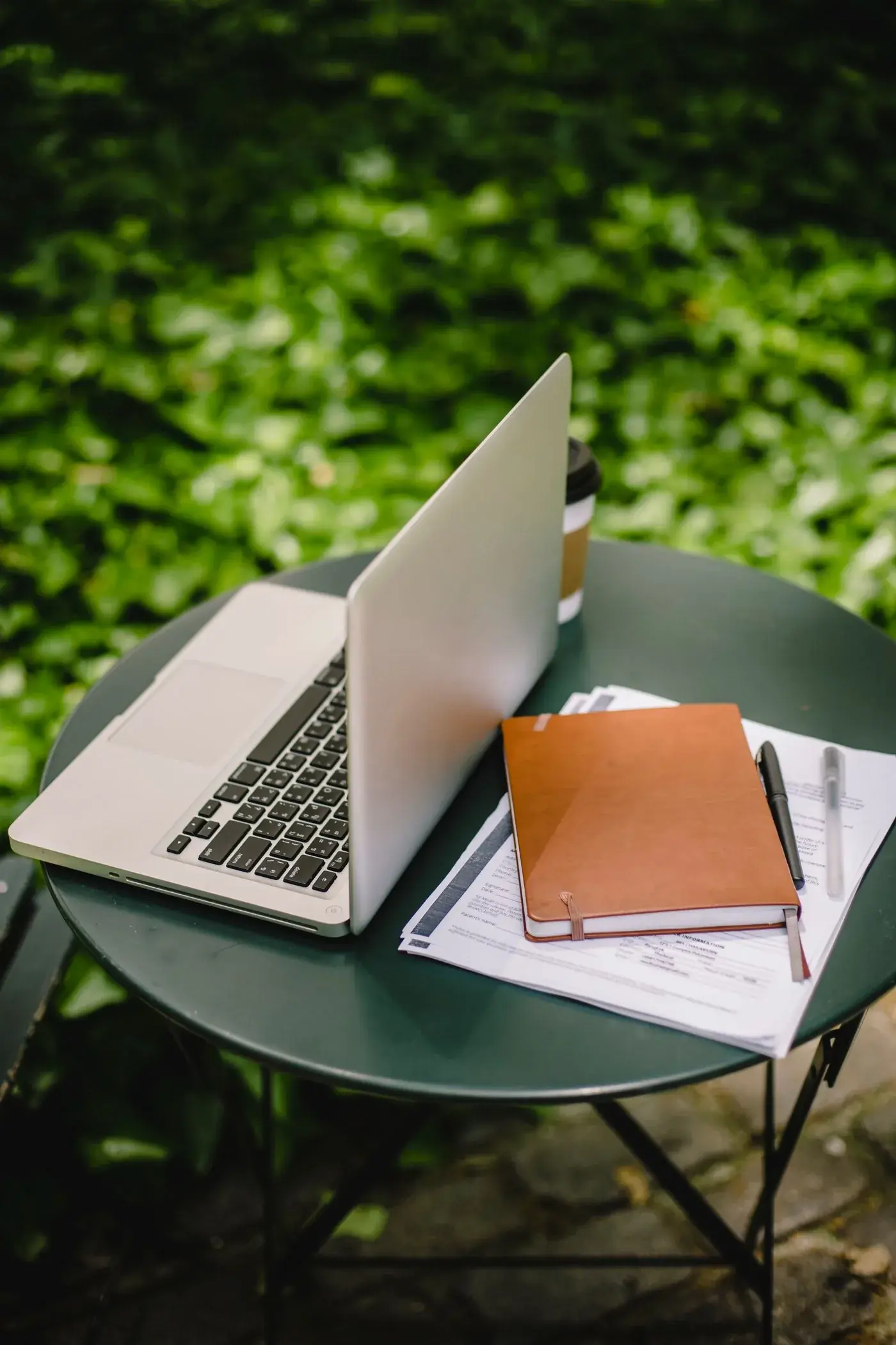 A laptop and notebook in a leafy outdoor workspace